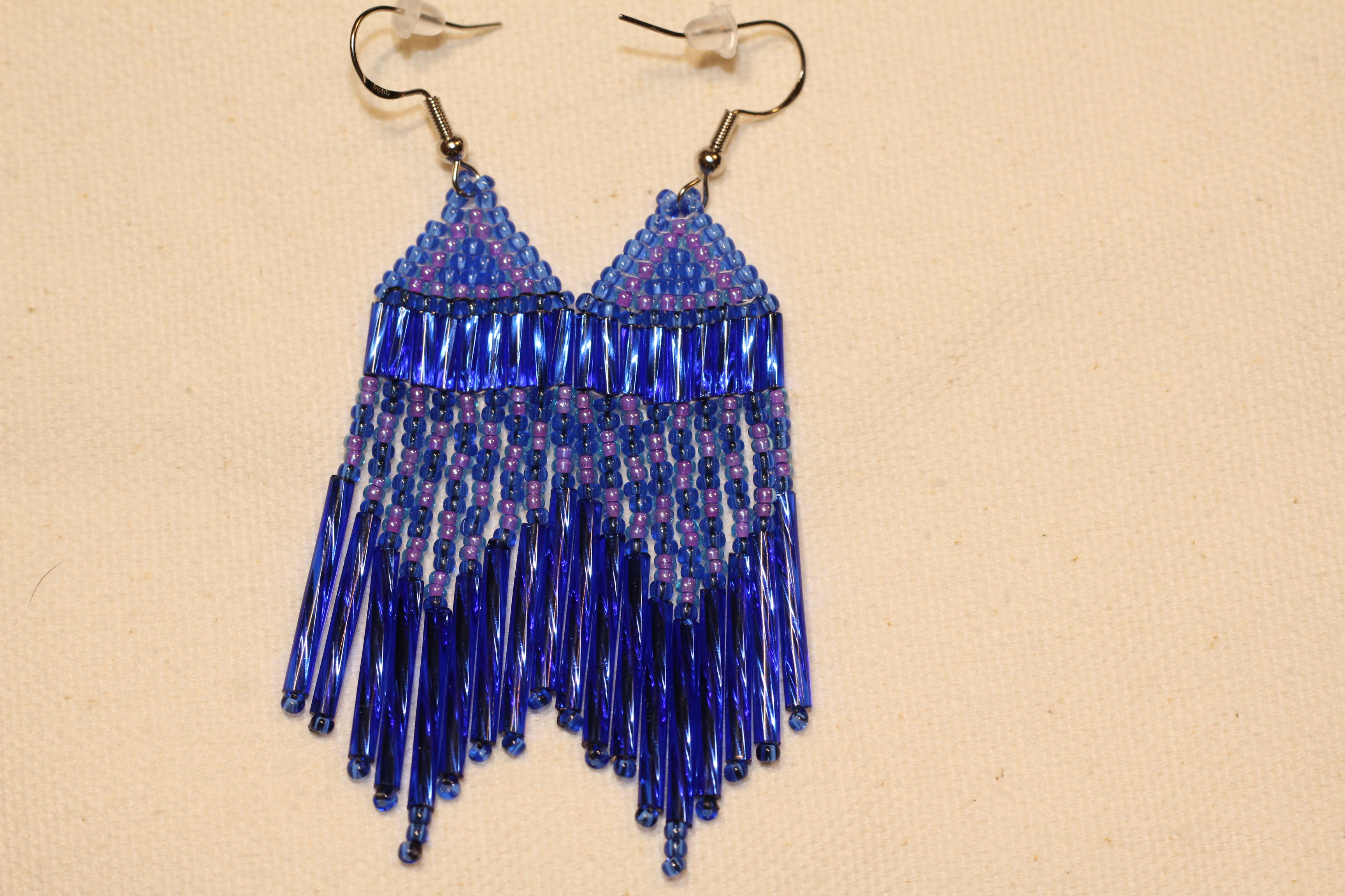 "Add a pop of shimmer with these unique blue and purple fringe earrings! Featuring a triangle pattern in transparent blue and shiny purple, the design is accented with royal blue bugle beads and a checkered fringe for extra sparkle. Handmade with the brick stitch technique, these lively earrings are perfect for anyone who loves playful, eye-catching accessories. Measurements: 0.70 inch wide, 3 inches long."